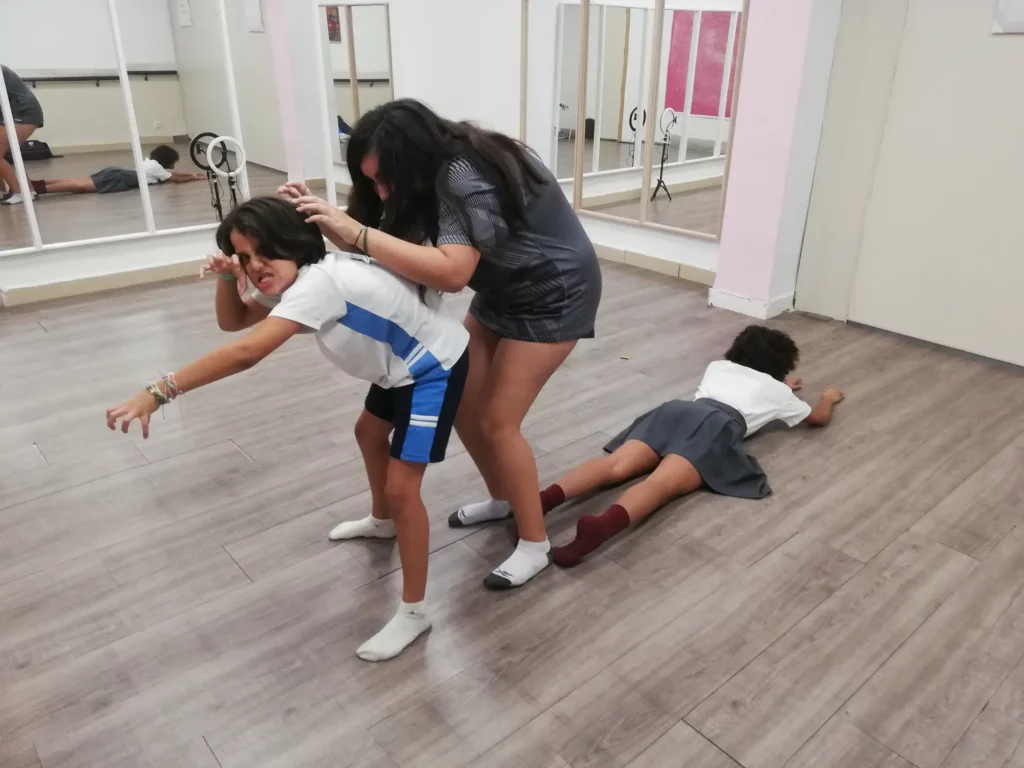 Three children are in a room with mirrored walls. One child lies face down on the floor, another child is standing and pushing their head down, and the third child stands behind the second, holding their hair. It looks like an intense drama class mimicking a school scene.